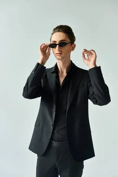 A young queer person strikes a confident pose in a black suit and sunglasses against a grey background. — Stock Photo