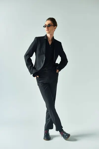 Homem gay jovem em um terno preto elegante marcando uma pose confiante e orgulhosa contra um fundo cinza. — Fotografia de Stock
