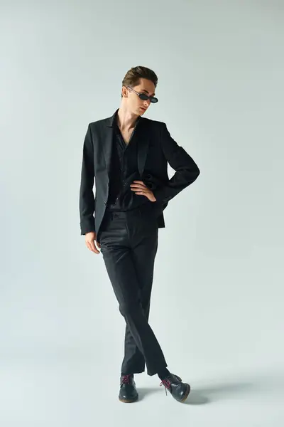 Uma jovem queer pessoa em um terno preto elegante marcando uma pose confiante em um estúdio contra um fundo cinza. — Fotografia de Stock