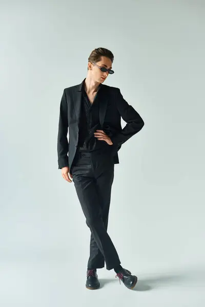 A young queer person in a sleek black suit striking a confident pose in a studio against a grey background. — Stock Photo