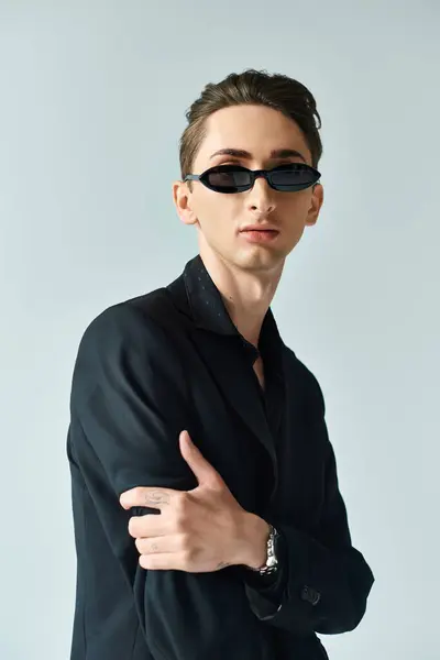 Une jeune queer en costume stylé et lunettes de soleil prend une pose confiante sur fond gris. — Photo de stock