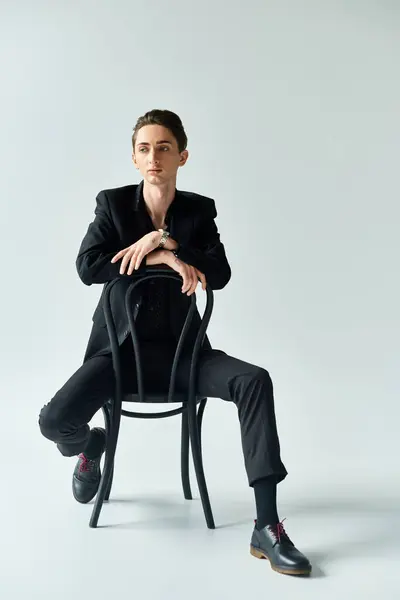 A young queer person confidently sits in a suit on a chair against a grey background, exuding pride and empowerment. — Stock Photo