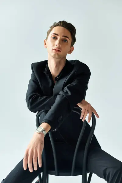 A young queer person exudes confidence and pride while sitting in a black suit on a chair in a studio against a grey background. — Stock Photo
