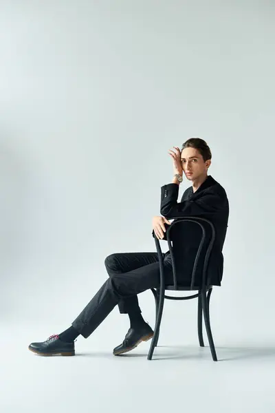 Un giovane elegante in giacca e cravatta si siede su una sedia, trasudando fiducia e contemplazione in uno studio con uno sfondo grigio. — Foto stock