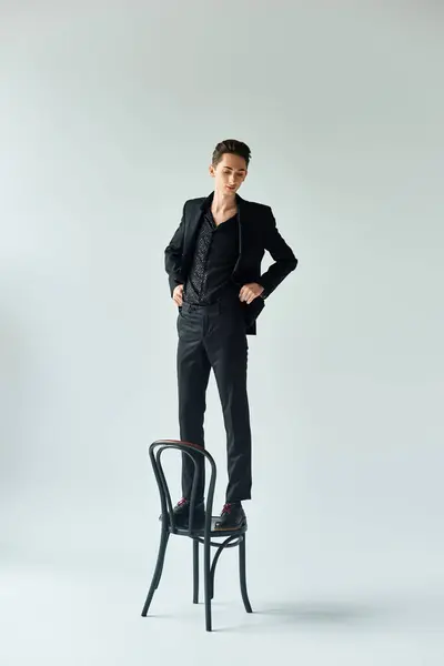 Hombre queer con estilo en un traje negro de pie con confianza en una silla en un entorno de estudio, mostrando orgullo y elegancia. - foto de stock