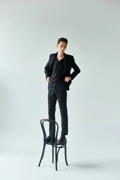Stylish queer man in a black suit standing confidently on a chair in a studio setting, showcasing pride and elegance. — Stock Photo
