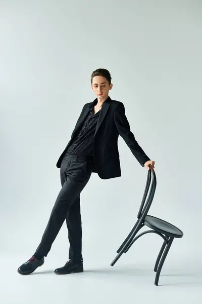 A young queer person in a black suit leans elegantly on a black chair, striking a confident and stylish pose in a studio. — Stock Photo