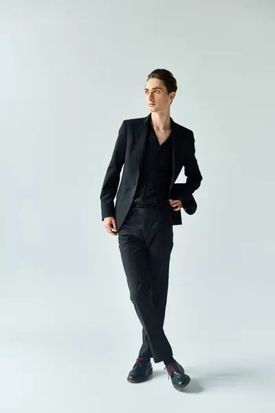 A young queer man, dressed in a sleek black suit, confidently poses for the camera in a studio with a grey background. — Stock Photo