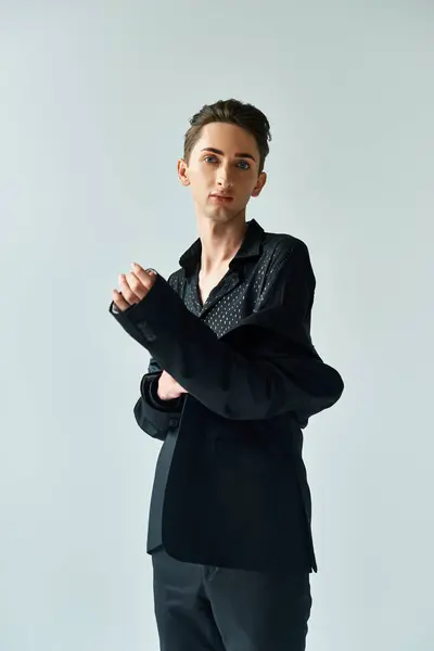 Un giovane uomo queer in un abito nero elegante colpisce una posa sicura su uno sfondo grigio studio. — Foto stock