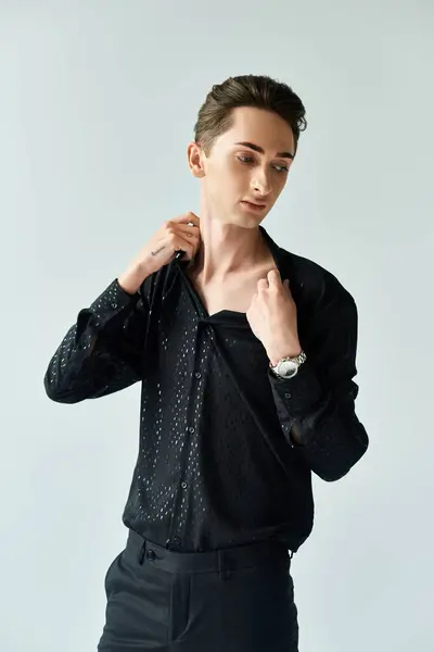 Un giovane uomo, parte della comunità LGBT, posa fiducioso in una camicia nera e pantaloni su uno sfondo grigio studio. — Foto stock