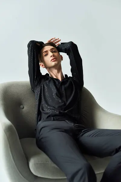 Un giovane omosessuale siede su una sedia, mani sopra la testa, profondamente pensieroso, su uno sfondo grigio da studio. — Foto stock