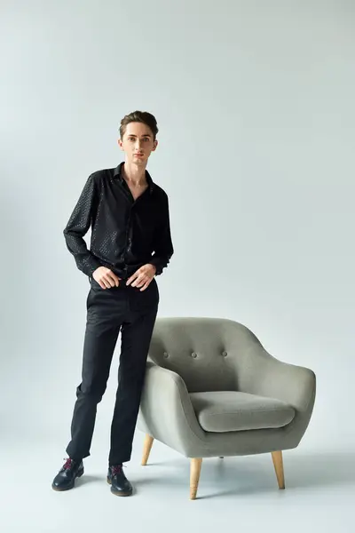 A young queer person in a black shirt striking a pose beside a sleek grey chair in a studio setting. — Stock Photo