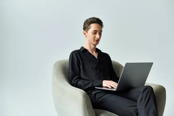 Ein junger queerer Mensch, der stolz seine Identität ausdrückt, sitzt auf einem Stuhl und benutzt einen Laptop in einem Studio mit grauem Hintergrund. — Stockfoto