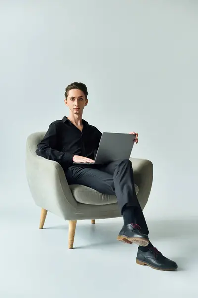 Un giovane omosessuale siede su una sedia con un computer portatile, profondamente assorto nel suo lavoro, su uno sfondo grigio da studio. — Foto stock