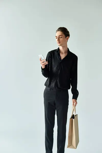 Un joven queer sosteniendo una bolsa de la compra, mira su teléfono sobre un fondo gris, mostrando multitarea en estilo urbano. — Stock Photo