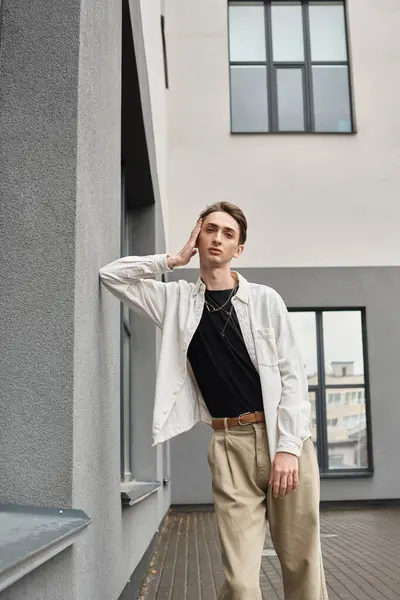 A young queer man leans against a wall in a stylish white shirt and beige pants, exuding confidence and pride. — Stock Photo