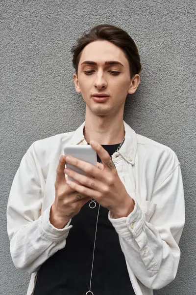 A young queer person in stylish attire leans against a wall, engrossed in his phone. — Stock Photo