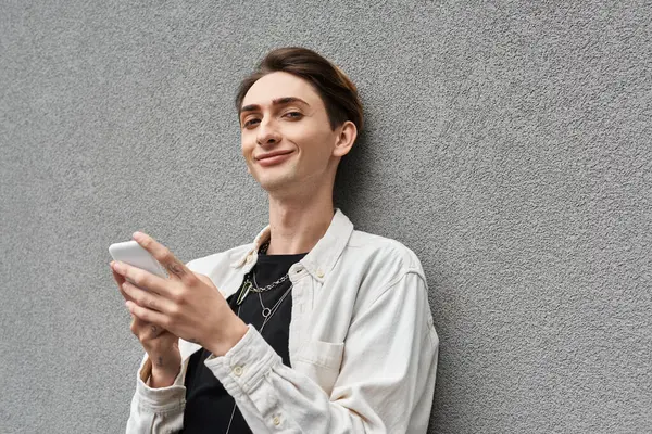 A young queer person in stylish attire leans against a wall, absorbed in his phone. — Stock Photo