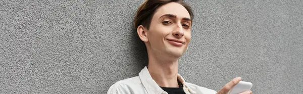 Ein stilvoll gekleideter junger queerer Mensch lehnt an einer Wand, in sein Handy vertieft. — Stockfoto