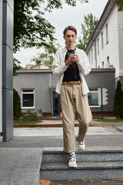 A young queer person in stylish tan jacket and khaki pants is standing on the steps, exuding confidence and pride. — Stock Photo