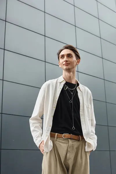 A young queer person in a white shirt and tan pants confidently poses in front of a building. — Stock Photo
