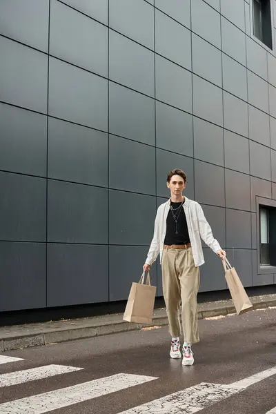 Стильна молода квір-людина ходить з торговими сумками перед елегантною міською будівлею, демонструючи впевненість і гордість. — стокове фото