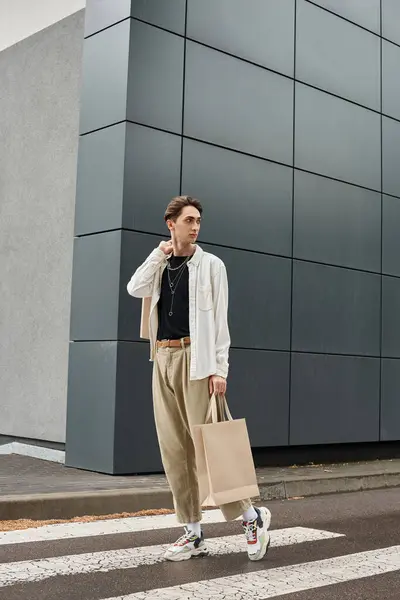 Um jovem gay em trajes elegantes cruza confiantemente a rua com um saco de compras na mão. — Fotografia de Stock