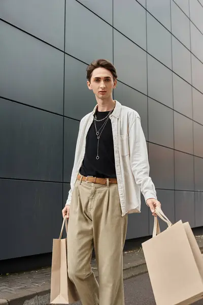 A young queer person in stylish attire, holding shopping bags, stands in front of a modern building. — Stock Photo