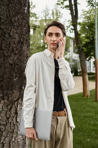 Um jovem, elegantemente vestido queer indivíduo de pé ao lado de uma árvore, conversando em seu telefone em um ambiente casual ao ar livre. — Fotografia de Stock