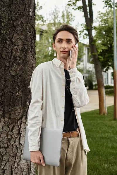 A young, stylishly dressed queer individual standing next to a tree, chatting on his phone in a casual outdoor setting. — Stock Photo