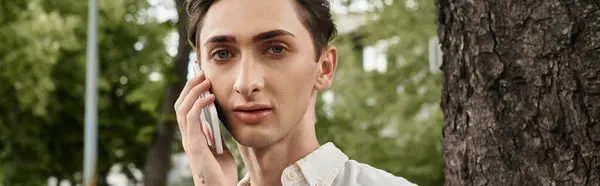 Un giovane individuo queer in abiti alla moda conversa su un cellulare di fronte a un albero, incarnando orgoglio e appartenenza. — Foto stock