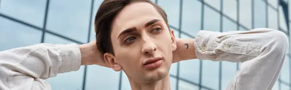 Un joven queer con un atuendo elegante mira hacia arriba, perdido en el pensamiento, enmarcado por el telón de fondo dinámico de un edificio moderno. — Stock Photo