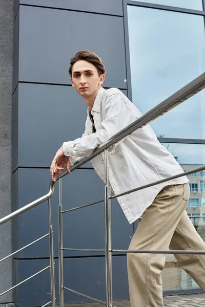 Un joven queer con un atuendo elegante se apoya pensativamente en una barandilla frente a un edificio, abrazando su identidad con orgullo. — Stock Photo