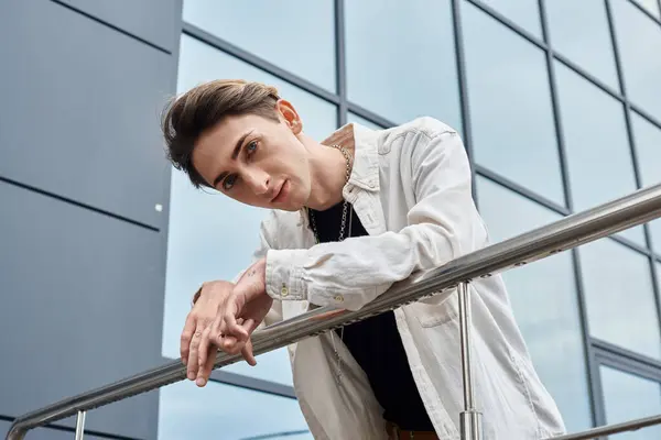 A vibrant young man, embodying queer pride, leans confidently on a railing in front of a modern building. — Stock Photo