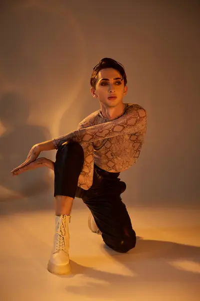 A young queer person, stylishly dressed, with tattoos, crouching in front of a bright light. — Stock Photo