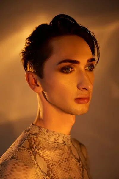 A young queer man in a snake skin dress stands gracefully in front of a bright light, exuding pride and style. — Stock Photo