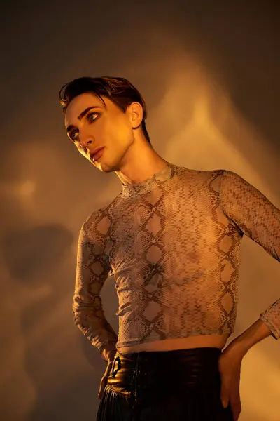 A young queer man confidently poses in front of a bright light in stylish top and leather pants. — Stock Photo
