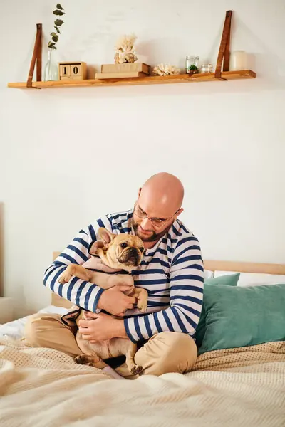 Man in glasses sitting on bed, embracing small french bulldog. — Stock Photo