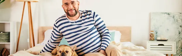 A man in glasses sits peacefully on a bed with his French bulldog beside him. — Stock Photo