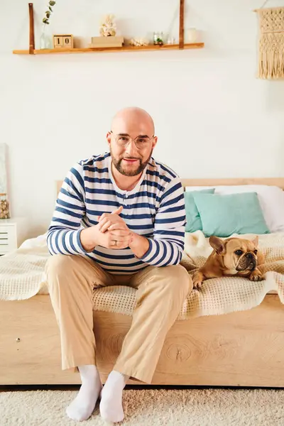Schöner Mann mit Brille sitzt auf einem Bett neben einer französischen Bulldogge. — Stockfoto