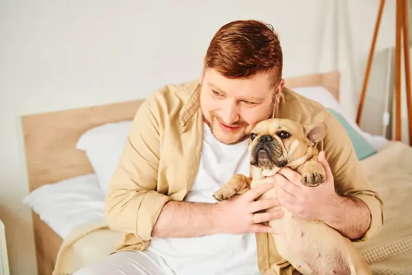 Ein stilvoller Mann sitzt auf einem Bett und hält liebevoll eine kleine französische Bulldogge. — Stockfoto