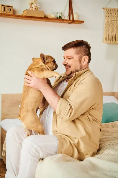 A man sitting on a bed, holding a French bulldog lovingly. — Stock Photo