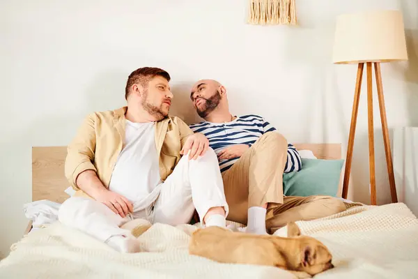An attractive gay couple sits comfortably on a bed with their French Bulldog. — Stock Photo
