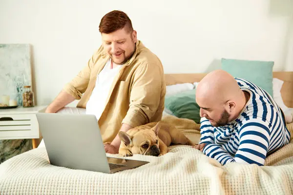 Dois homens e um cão descansando em uma cama enquanto usando um laptop. — Fotografia de Stock