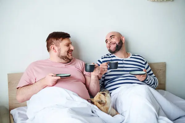 A gay couple relaxes on a bed with their French bulldog nearby. — Stock Photo