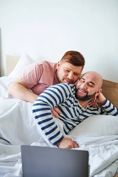 Dos hombres se acomodan en la cama mientras usan un portátil. - foto de stock