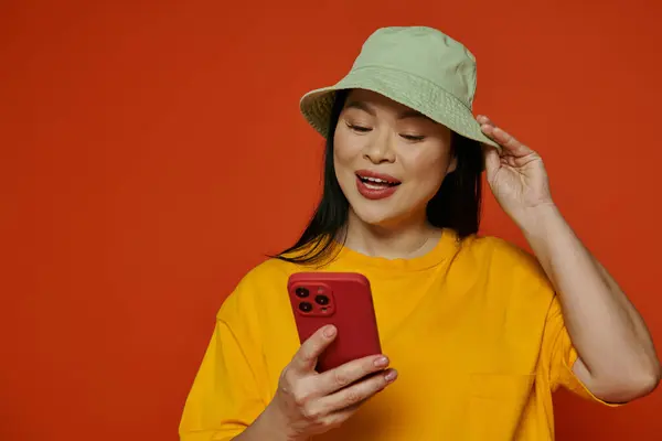 Une femme asiatique à la mode dans un chapeau tenant un téléphone portable sur un fond orange. — Photo de stock