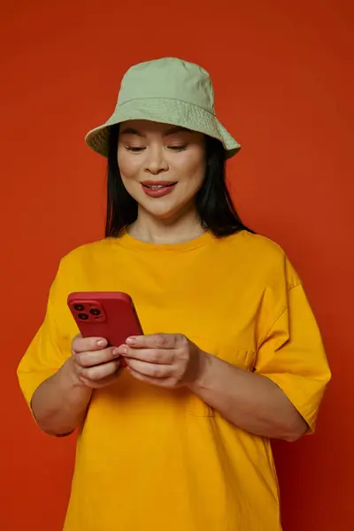 Une femme dans un chapeau parcourant son téléphone portable dans un studio avec une toile de fond orange. — Photo de stock