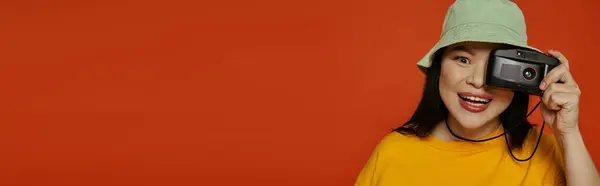 An Asian woman holds a camera to her face in a studio against an orange backdrop. — Stock Photo