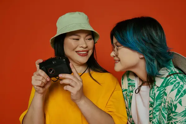 Asian mother and teenage daughter standing together, holding a camera in a studio with an orange background. — Stock Photo